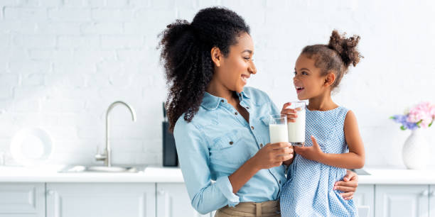 uśmiechnięta afroamerykańska matka i córka trzymające szklanki mleka w kuchni i patrzące na siebie - preschooler child offspring purity zdjęcia i obrazy z banku zdjęć