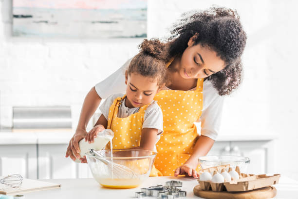 afro-américaine mère et fille prépare la pâte et verser le lait dans bol dans la cuisine - preschooler child offspring purity photos et images de collection