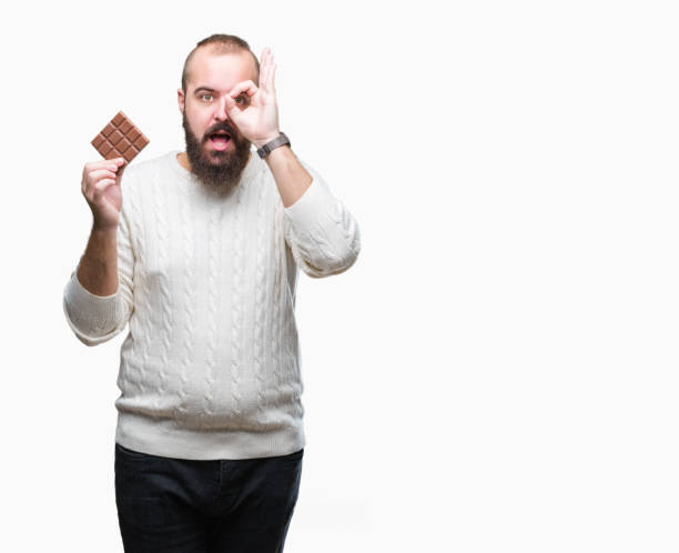 istock Young hipster man eating chocolate bar over isolated background with happy face smiling doing ok sign with hand on eye looking through fingers 1067799470