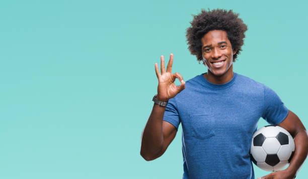 афро американский человек, держащий футбольный мяч над изолированным фоном делать нормально знак пальцами, отличный символ - football human hand holding american football стоковые фото и изображения