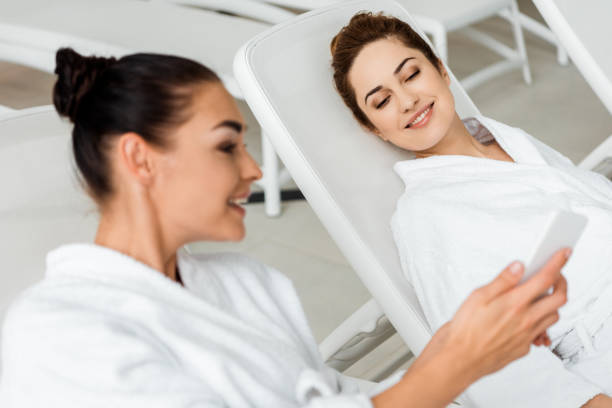 istock smiling young women in bathrobes using smartphone and talking while relaxing in spa 1067809326