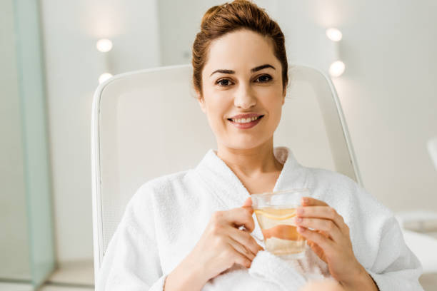 istock young woman in bathrobe holding cup with herbal drink and smiling at camera while relaxing in spa 1067814274
