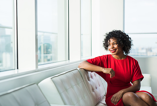 A portrait of happy black woman sitting on a sofa by the window in an office. Copy space.
