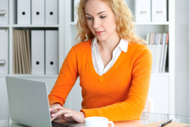 istock Young beautiful business woman or student communicates by laptop computer while sitting at the desk and holding black pen. She Orange sweater is at the girl. Education, business and lifestyle concept. 1067831144