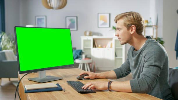 istock Handsome Young Man Works on a Green Mock-up Screen Personal Computer while Sitting at His Desk in the Cozy Apartment. 1067853994