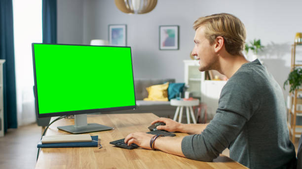 istock Handsome Young Man Works on a Green Mock-up Screen Personal Computer while Sitting at His Desk in the Cozy Apartment. 1067854020