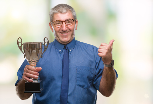 Handsome senior successful man holding trophy over isolated background pointing and showing with thumb up to the side with happy face smiling
