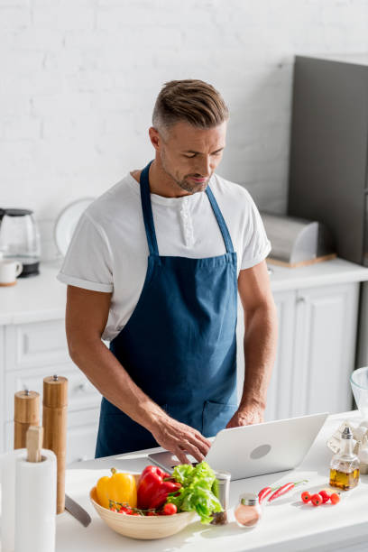 istock handsome man in apron standing at kitchen and working on laptop 1067884972