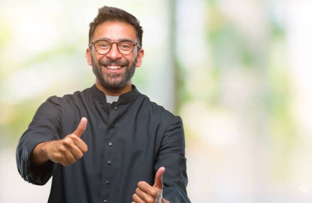 istock Adult hispanic catholic priest man over isolated background approving doing positive gesture with hand, thumbs up smiling and happy for success. Looking at the camera, winner gesture. 1067907940
