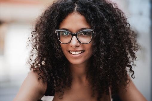 Close-up outdoor portrait of a dazzling young caucasian woman with curly bulky hair, brown eyes and in eyeglasses, she is smiling and looking at the camera, shallow depth of field
