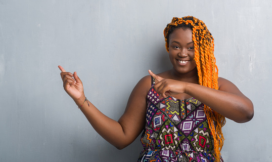 Young african american woman over grey grunge wall wearing orange braids smiling and looking at the camera pointing with two hands and fingers to the side.