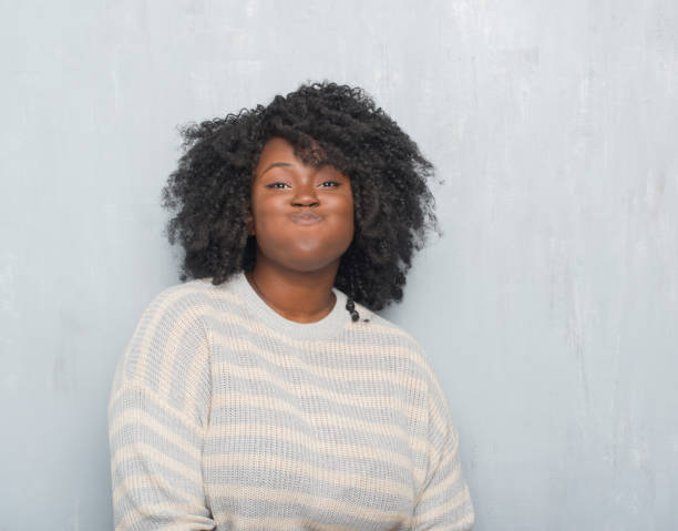 jóvenes afroamericanos más mujer de tamaño sobre la pared gris grunge usando un suéter soplar las mejillas con cara divertida. boca se infla con aire, expresión loco. - chubby cheeks fotografías e imágenes de stock