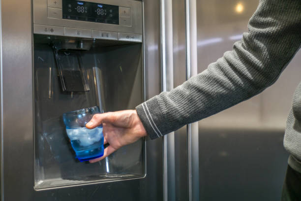 la mano maschile versa acqua fredda e cubetti di ghiaccio dal distributore del frigorifero di casa. - ice machine foto e immagini stock