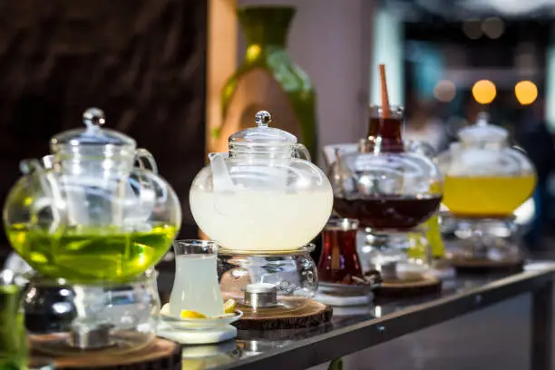 Close up color image depicting a variety of exotic and fruit teas for sale at an outdoor food and drink market. The tea - including lemon and ginger, and mint tea - is being infused inside clear glass teapots on the bar counter. The serving staff are defocused in the background.