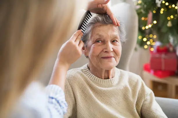 An unrecognizable health visitor combing hair of senior woman sitting on a sofa at home at Christmas time.