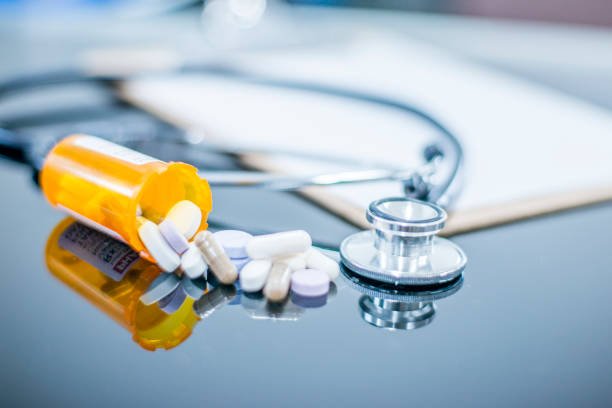 Bottle of pills spills onto doctor's desk A bottle of pills is tipped on its side, spilling onto a glass-top desk with stethoscope and clip board in the background. opioid dependency stock pictures, royalty-free photos & images