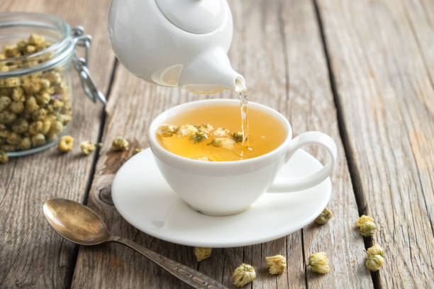 té de manzanilla saludable vierte en taza blanca. tetera y cuchara, tarro de cristal de las hierbas medicinales de daisy. - manzanilla fotografías e imágenes de stock