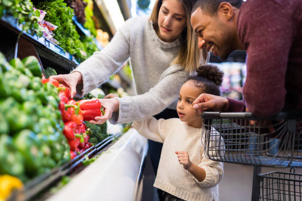 A pre-school age girl helps her parents pick out veggies in the produce section at the grocery store. She is reaching for a red pepper.
