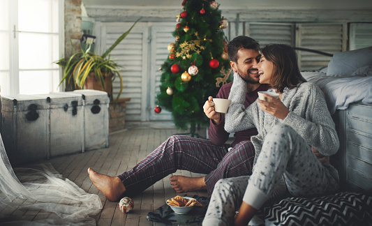 Closeup of a mid 20's couple having some coffee and relaxing by a window on a Christmas morning. They are laughing and captured in a very spontaneous moment.