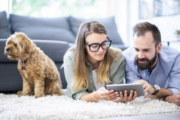 Couple laying on a rug looking at a tablet A caucasian couple lies on their living room rug, with their dog in the background, looking at a tablet. borrowing stock pictures, royalty-free photos & images
