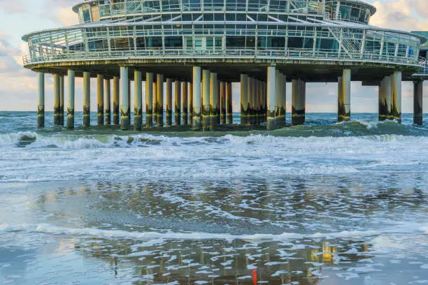 Photo of building on stone weathered poles at the beach of scheveningen with waves in the sea and reflection in the wet sand