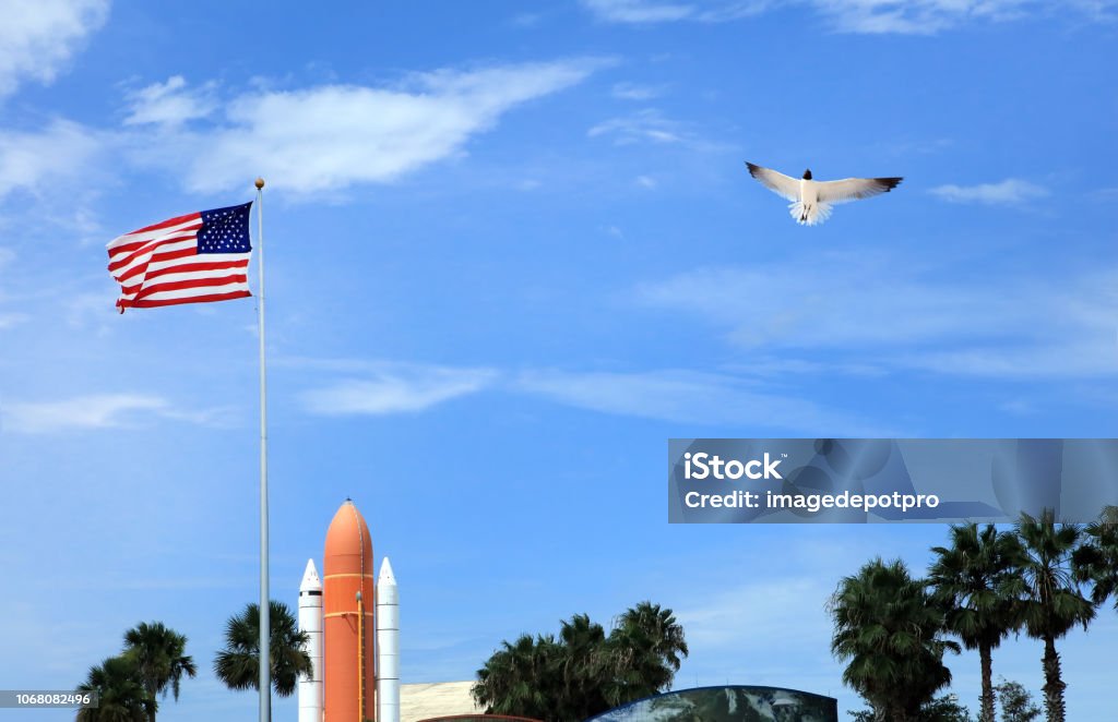 Waving American flag at Kennedy space center entrance with space rocket Waving American flag at Kennedy space center entrance with space rocket and palm trees over blue sky in Florida, USA Admiration Stock Photo