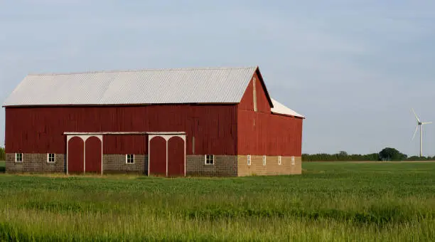 An old wooden farm barn painted red with at new electricity producing wind turbine in the distance.