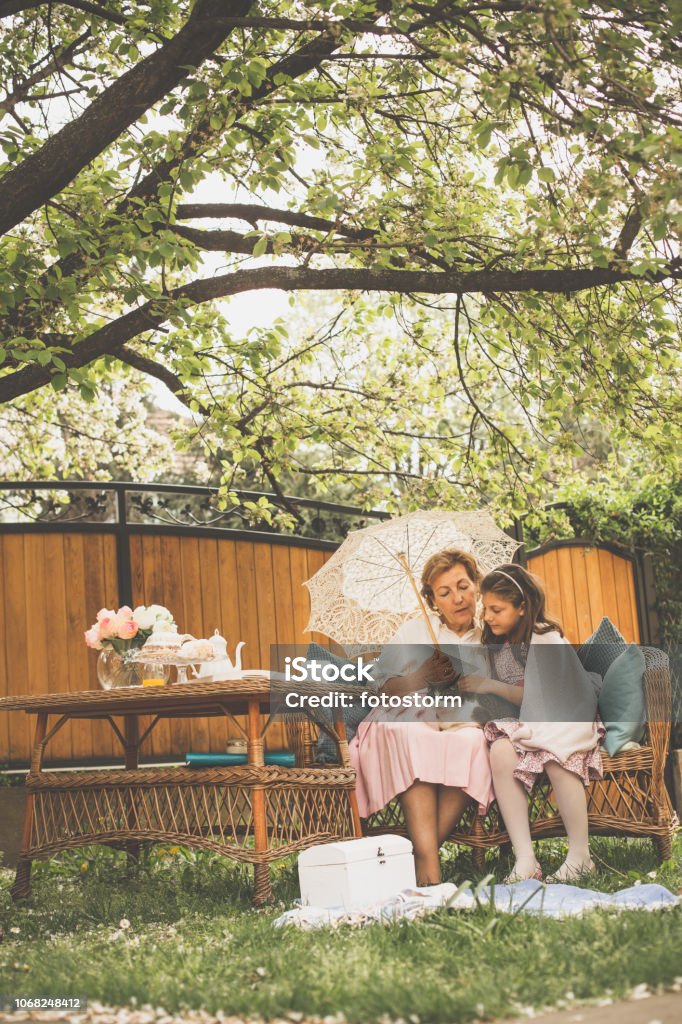 Grandmother and granddaughter relaxing in the garden A senior woman and her granddaughter are relaxing and cuddling a cat in the back yard on a beautiful spring day. The woman is holding a vintage parasol. Grandchild Stock Photo