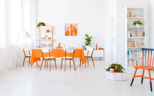 istock Bright dining room interior with window with drapes, rack with books, fresh plants and table with orange tablecloth, four chairs and dinnerware in the real photo 1068259168