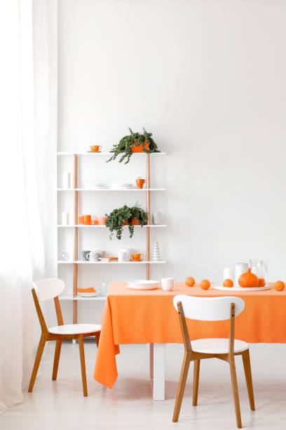 istock Real photo of white dining room interior with empty wall, rack with plants, cups and plates, wooden chairs and orange cloth on table with dinnerware 1068259172