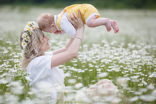 A happy mother holds in her arms her newborn son standing outdoors in the highlands among a blooming chamomile field. Family, mother and baby, against the backdrop of mountains and blue sky on a white meadow of blooming daisies