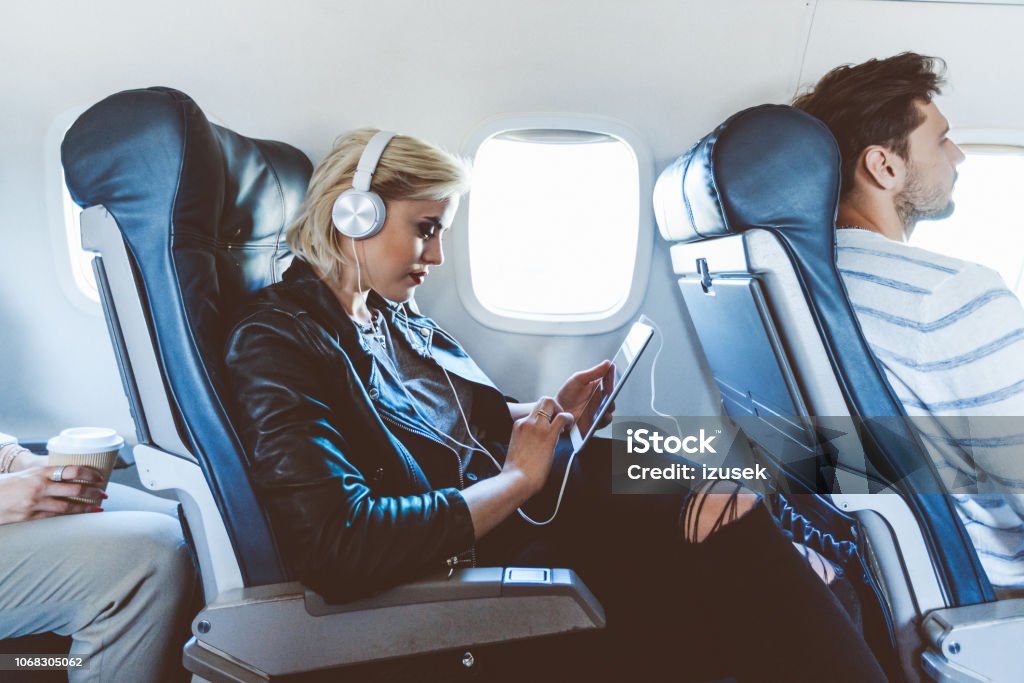 Female passenger using digital tablet during flight Young blonde woman sitting inside an airplane and using a digital tablet. Female passenger using e-reader during flight. Airplane Stock Photo