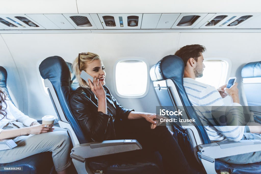 Female passenger talking on mobile during flight Young blonde woman sitting inside an airplane and talking on mobile phone. Adult Stock Photo
