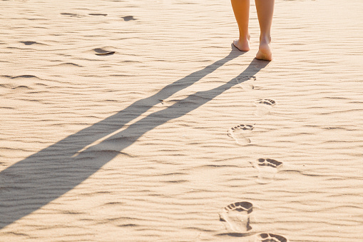 Young woman's barefoot walking on the dry sand in sunny summer day. Part of body. Go forward. Back view. Long shadow. Leaving footprints.