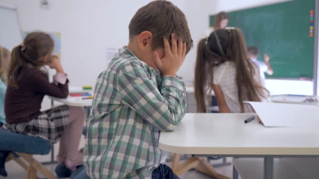 bullying, tired schoolboy sits at a desk in classroom on background of classmates and female teacher near blackboard