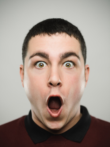 Close-up portrait of a man with the mouth wide open and a shocked expression looking at camera. Young caucasian male with brown hair against white background. Vertical studio photography from a DSLR camera. Sharp focus on eyes.