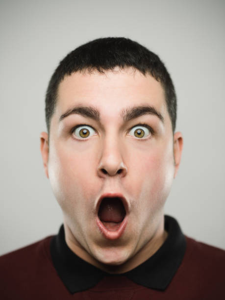 Portrait of a surprised young caucassian man looking at camera. Close-up portrait of a man with the mouth wide open and a shocked expression looking at camera. Young caucasian male with brown hair against white background. Vertical studio photography from a DSLR camera. Sharp focus on eyes. mouth open human face shouting screaming stock pictures, royalty-free photos & images