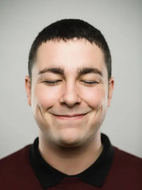 Close-up portrait of real young man with closed eyes and with a comfortable attitude. Caucasian male has brown hair. He is against gray background. Vertical studio photography from a DSLR camera. Sharp focus on eyes.