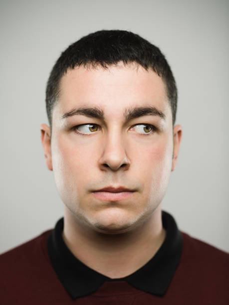 Portrait of a real young caucassian man looking away. Close-up portrait of real young man looking around with blank expression. Serious caucasian male with brown hair against gray background. Vertical studio photography from a DSLR camera. Sharp focus on eyes. sideways glance stock pictures, royalty-free photos & images