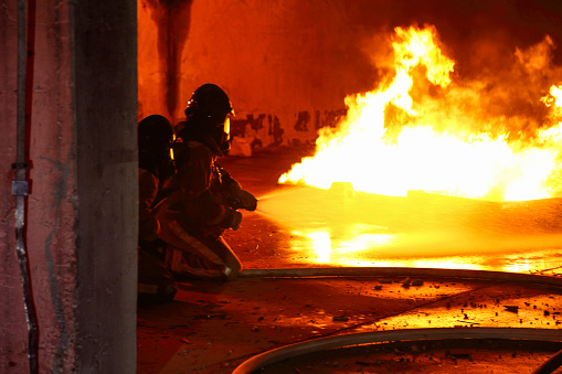 Firemen spraying water onto a big fire. Two firefighters kneeling near the flames in a protective suits and helmets.