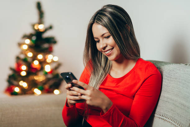 istock Young woman with mobile phone at home while sitting on sofa near Christmas tree 1068340818