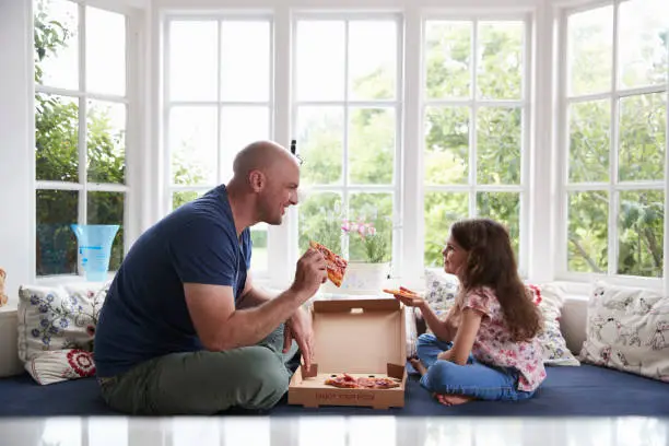 Photo of Dad and daughter sit on window seat at home sharing a pizza