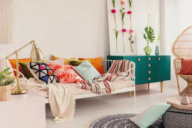 Photo of Patterned and colorful pillows on single metal bed in stylish girl's bedroom interior with peacock chair and green cabinet with crown shape handles