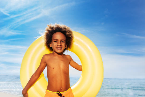 Close-up portrait of African boy holding big swim ring behind his back, standing against exotic seascape and looking at camera
