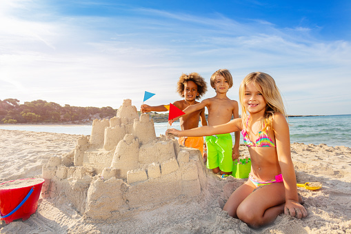 Happy friends, multiethnic boys and girls, putting flags in sandcastle towers, playing beach games in summer