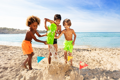 Portrait of multiethnic boys, happy friends, making sandcastle together on the beach