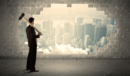 Business man hitting wall with hammer on city view background