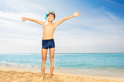 Full-length portrait of preteen boy in scuba mask standing on the beach with his hands up against tropical seascape in summer