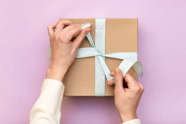 woman hands holding craft gift box with blue bow on pastel pink background. girl binds classic bow for present box. top view, flat lay style, copy space - hand wrap imagens e fotografias de stock