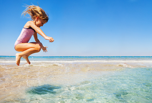 Side view portrait of joyful little girl in bikini jumping at shallow water against beautiful seascape background